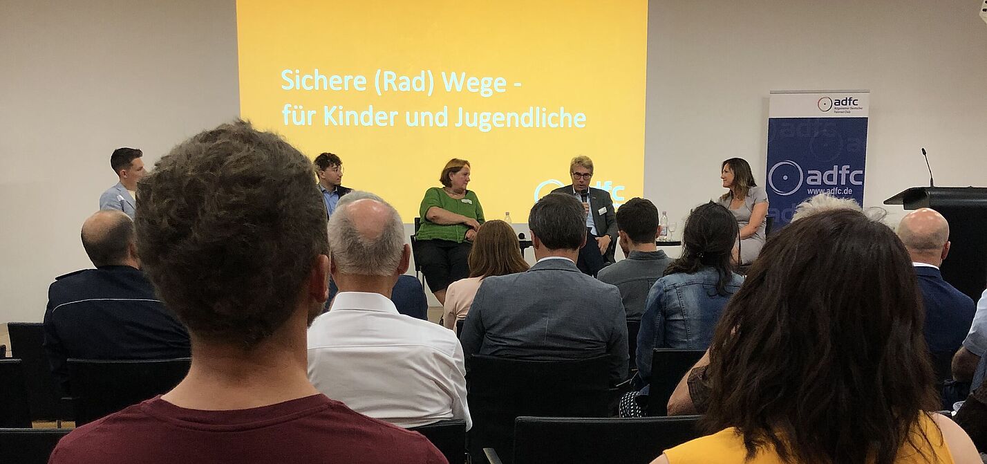 Bild der Podiumsdiskussion des ADFC. Im Hintergrund ist die Bühne mit den Diskussionsteilnehmenden und dem Thema "Sichere (Rad)Wege für Kinder und Jugendliche" zu sehen. Vorne sind Hinterköpfe des Publikums zu sehen © ADFC Baden-Württemberg